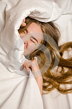 Beautiful young woman wakes up in the morning in white bed in the rays of the sun, she has lush wavy blonde, golden hair