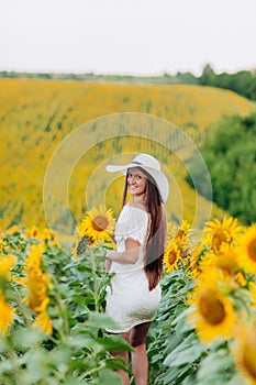 Beautiful young woman walking in blooming sunflowers field in summer. Stylish girl with long hair in white dress and hat. summer