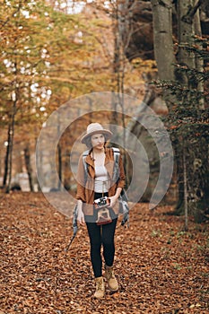 Attractive young woman in a stylish hat and travel bag on her shoulders, looking around at the charming autumn forest