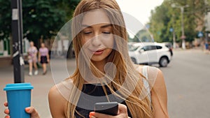 Beautiful Young Woman Walking in the City. Using her Mobile Phone. Typing a Message. Smiling Happily. Drinking Delicious