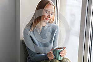 Beautiful young woman in warm sweater drinking hot tea on windowsill at home