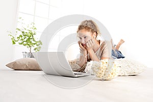 Hermoso mujer joven mirar una película sobre el computadora comer maiz tostado acostada sobre el sala de estar de madera piso en cómodo 