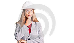 Beautiful young woman wearing architect hardhat with hand on chin thinking about question, pensive expression