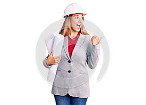 Beautiful young woman wearing architect hardhat holding build project pointing thumb up to the side smiling happy with open mouth