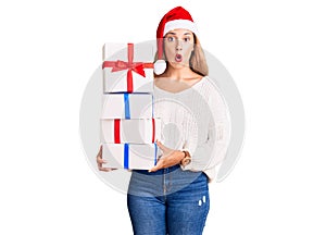 Beautiful young woman wearing christmas hat and holding a gift scared and amazed with open mouth for surprise, disbelief face