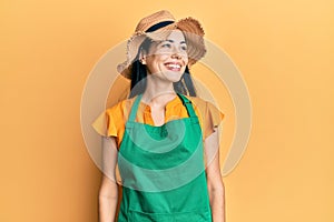 Beautiful young woman wearing gardener apron and straw hat looking to side, relax profile pose with natural face and confident