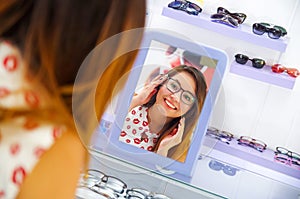 Beautiful young woman wearing glasses in optical store and looking in the mirror how she looks, in a blurred background