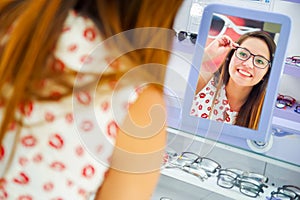 Beautiful young woman wearing glasses in optical store and looking in the mirror how she looks, in a blurred background