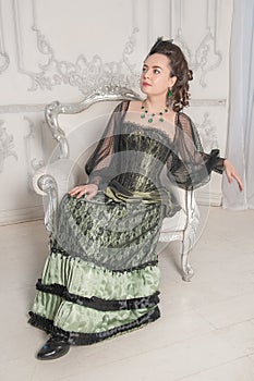 Beautiful young woman wearing green medieval vintage Victorian Style dress sitting in the armchair