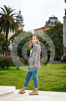 Beautiful young woman wearing jacket and jeans walking on the sidewalk. Palm trees and historic building facade on