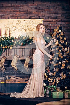 A beautiful young woman wearing a long Golden dress adorns the Christmas tree with toys and balls in the house near the fireplace