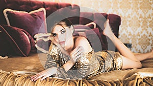 Beautiful young woman wearing luxury glitter dress lying in bed and relaxing. Rich interior style. Sensual attractive