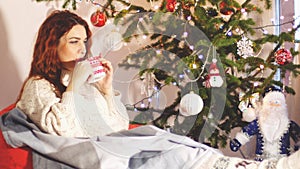 Beautiful young woman wearing stylish christmas sweater and socks sitting on comfortable sofa and drinks tea by