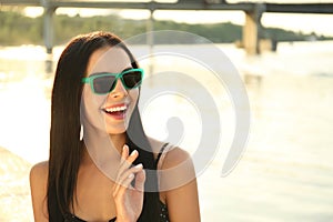 Beautiful young woman wearing stylish sunglasses near river. Space for text