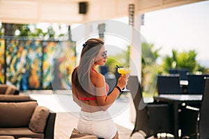 Beautiful young woman wearing swimsuit drinking a colorful cocktail sitting on a cabin of the beach club bar. Stunning