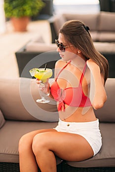 Beautiful young woman wearing swimsuit drinking a colorful cocktail sitting on a cabin of the beach club bar. Stunning