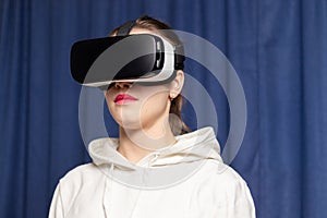 Beautiful young woman wearing virtual reality glasses and a white hoodie on a blue background. Modern technologies. Selective focu