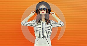 Beautiful young woman wearing a white striped shirt, black round hat on orange background