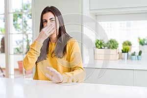 Beautiful young woman wearing yellow sweater smelling something stinky and disgusting, intolerable smell, holding breath with