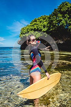 beautiful young woman in wetsuit and sunglasses with surfboard posing in ocean at Nusa dua Beach