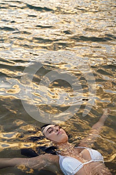 Beautiful young woman in white bikini laying down in the sea water. Sunset golden light. Calm, serene people