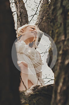 A beautiful young woman in a white chiffon dress with a short blonde haircut is sitting on a tree, bottom view