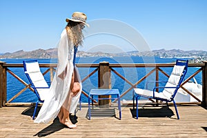 Beautiful young woman in white dress and bikini straw hat on white terrace balcony of house or hotel with Sea View