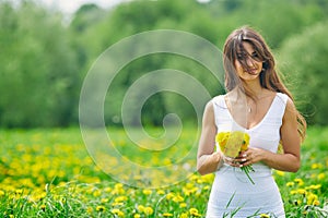 Beautiful young woman in a white dress collects dandelions on a green meadow