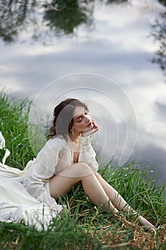 Beautiful young woman in white dress resting on shore lake pond river. Professional makeup and hairstyle with curled hair