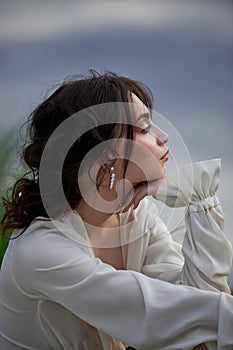 Beautiful young woman in white dress resting on shore lake pond river. Professional makeup and hairstyle with curled hair