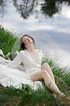 Beautiful young woman in white dress resting on shore lake pond river. Professional makeup and hairstyle with curled hair