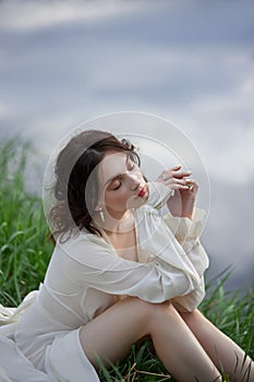 Beautiful young woman in white dress resting on shore lake pond river. Professional makeup and hairstyle with curled hair