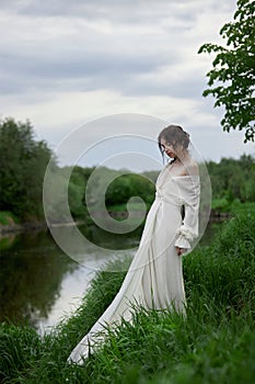 Beautiful young woman in white dress resting on shore lake pond river. Professional makeup and hairstyle with curled hair