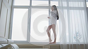 Beautiful young woman in a white men`s shirt and socks walking on windowsill, laughing and talking on the phone happily