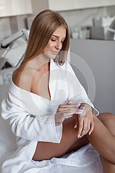 Beautiful young woman in a white robe applies moisturizing cream to her hands