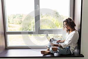 Beautiful young woman in white shirt, blue jeans sitting on windowsill laptop computer