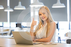 Beautiful young woman in white shirt is working on laptop and smiling while sitting in work place