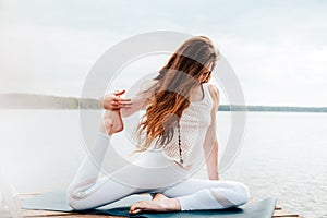 Beautiful young woman in white sports clothes is sitting on  wooden pier on lake in  lotus position with namaste gesture