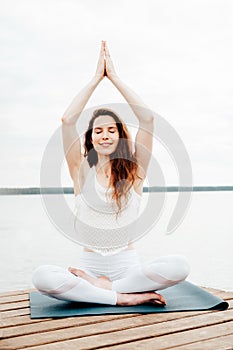 Beautiful young woman in white sports clothes is sitting on  wooden pier on lake in  lotus position with namaste gesture