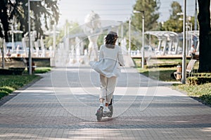 Beautiful young woman in white suit is riding to work on her electric scooter in city parkland