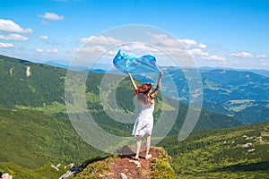 Beautiful young woman in white summer dress stands on a rock and looking into the amazing valley, blue scarf waving on the wind in