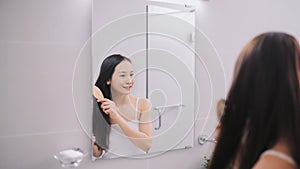 Beautiful young woman in white undershirt is combing her hair and smiling while looking into the mirror in bathroom