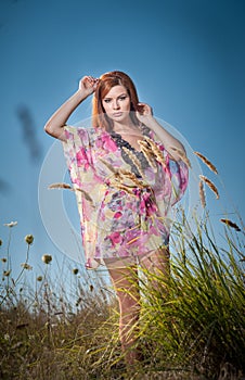 Beautiful young woman in wild flowers field on blue sky background. Portrait of attractive red hair girl with long hair relaxing