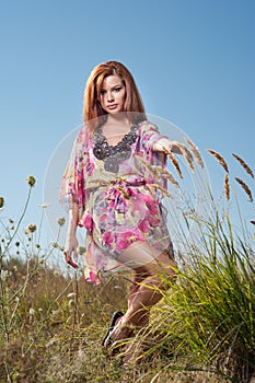 Beautiful young woman in wild flowers field on blue sky background. Portrait of attractive red hair girl with long hair relaxing