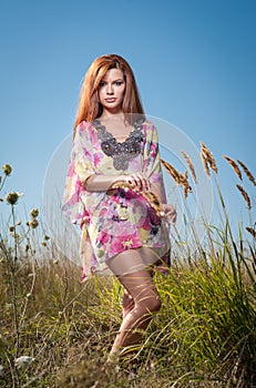 Beautiful young woman in wild flowers field on blue sky background. Portrait of attractive red hair girl with long hair relaxing