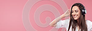 Beautiful young woman in wireless headphones listening to music and dancing on pink background.