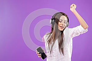 Beautiful young woman in wireless headphones listening to music using mobile phone and dancing on purple background