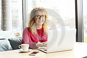 Beautiful young woman working remotely on her fashionable laptop at hipster coffee shop. Happy female freelancer with trendy wrist