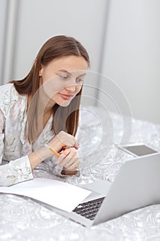 Beautiful young woman working, using laptop computer and makes notes in the bedroom at home. Freelancer. Writing, typing. Girl