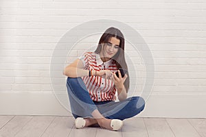 Beautiful young woman writing a text on her mobile phone while sitting cross-legged, isolated white brick wall background. One